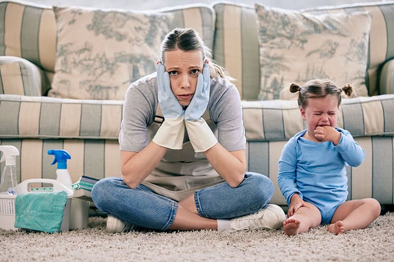 woman sitting on a rug wearing cleaning gloves looks defeated while toddler cries beside her