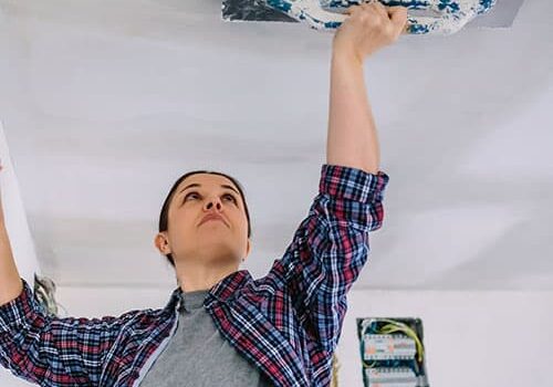 woman in a plaid shirt mudding drywal on a ceiling